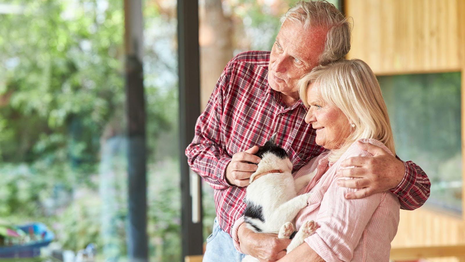 An elderly couple sharing a tender moment in their home, embracing each other with a smile while holding a black and white cat.