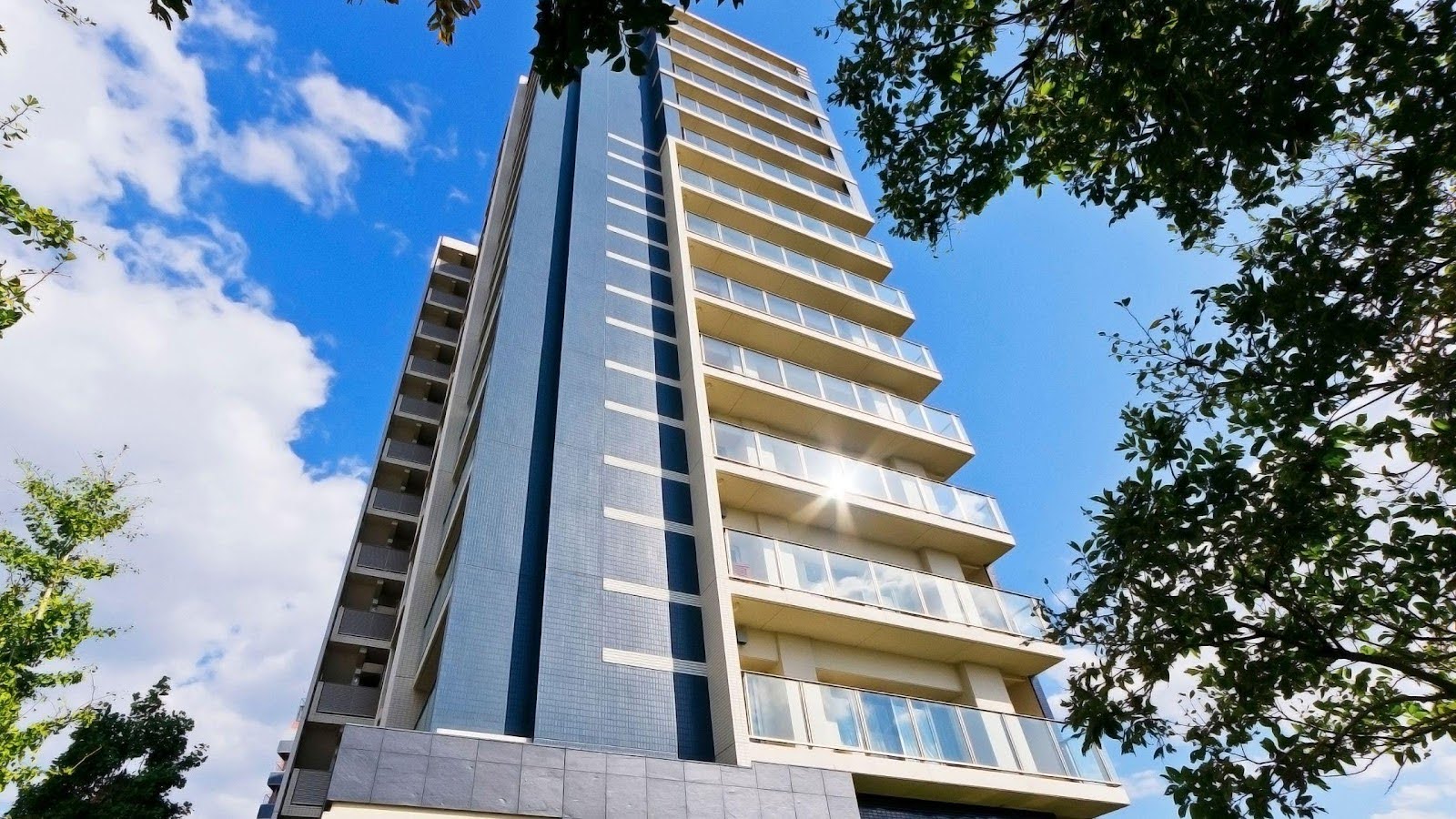 Modern high-rise apartment building viewed from a low angle, showcasing its towering structure and contemporary design with multiple balconies against a clear blue sky, framed by green tree branches at the top.